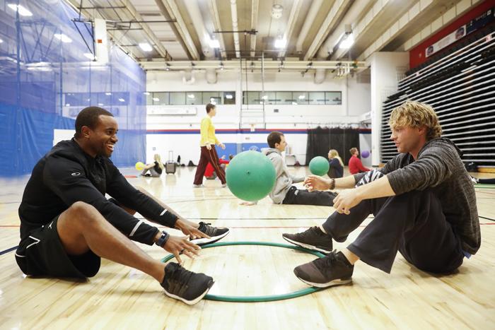 Photo of two adult students sitting on the floor in a gym with a hula hoop on the floor between them and children in the background.