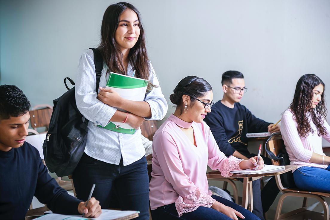 Students in class, one standing with a backpack and notebook
