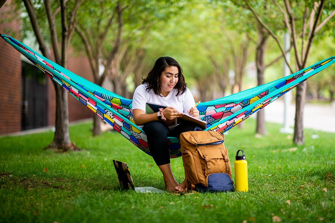Student sitting in hammock between trees on Auraria campus, with notebook in lap and laptop water bottle and backpack on ground next to them